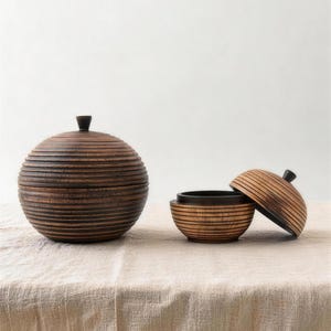 Könnte beinhalten: Zwei dekorative Holzschalen mit Deckeln. Die größere Schale ist rund mit dunkler brauner Oberfläche und einem kleinen Knopf oben. Die kleinere Schale ist offen und zeigt ein schwarzes Innere, der Deckel ist teilweise abgenommen. Beide Schalen haben ein Streifendesign.