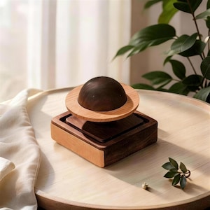 Pode incluir: Um objeto decorativo de madeira que se assemelha a um planeta. Apresenta um topo em forma de cúpula castanho escuro com um anel castanho claro, colocado sobre uma base quadrada de madeira de duas cores. A base é uma combinação de madeira castanha clara e escura.