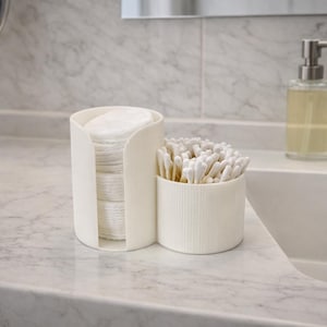 Könnte beinhalten: Ein weißer, geriffelter Badezimmer-Organizer mit zwei Fächern für Wattepads und Wattestäbchen. Der Organizer steht auf einer Marmorarbeitsplatte neben einem Waschbecken, einem Seifenspender und einem Wasserhahn. Das Design ist minimalistisch und modern.