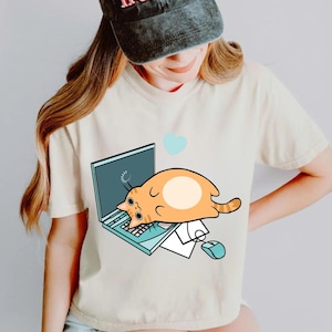 Könnte beinhalten: Cremefarbenes T-Shirt mit einer Cartoon-Katze, die auf einem Laptop liegt. Die Katze ist orange mit einem weißen Bauch. Ein blaues Herz schwebt über dem Laptop. Eine Computermaus und Papiere sind ebenfalls sichtbar.