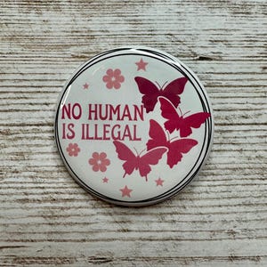 May include: A round button with a white background, black border, and the text "NO HUMAN IS ILLEGAL" in red. The button features pink butterflies, flowers, and stars.