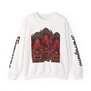 Op de afbeelding: Witte sweatshirt met een afbeelding van drie rode en gouden Japanse demonenmaskers. De mouwen hebben zwarte tekst. Een crew neck sweatshirt met een opvallend ontwerp.