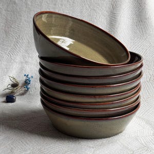 Może przedstawiać: Stos siedmiu ceramicznych misek z błyszczącym, przygaszonym oliwkowozielonym wykończeniem wewnątrz i na zewnątrz. Każda miska ma wyraźną, ciemną czerwonobrązową krawędź, która kontrastuje z ziemistą zieloną glazurą, tworząc jednolity zestaw naczyń.