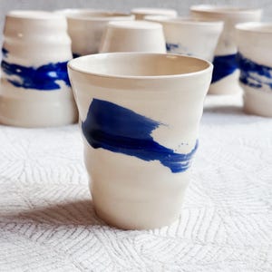 Puede incluir: Conjunto de tazas de cerámica hechas a mano. Cada taza es de color blanco roto con un diseño de pincelada azul intenso. Las tazas tienen una forma ligeramente irregular, lo que añade atractivo artesanal. Están dispuestas sobre una superficie blanca texturizada.