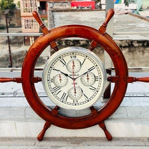 Könnte beinhalten: Eine Uhr in Form eines Holzschiffsrads mit einem weißen Zifferblatt und römischen Ziffern. Die Uhr hat drei kleinere Zifferblätter und die Worte "WORLD TIME" sind auf dem Zifferblatt aufgedruckt. Das Rad ist dunkelbraun.