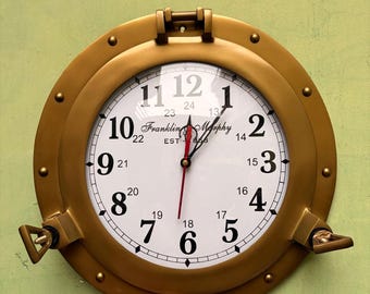 May include: A round brass-colored wall clock designed like a ship porthole. The white clock face features black Arabic numerals from 1 to 12, smaller 13 to 24 hour markers, and the text Franklin Morphy EST 1863. It has black hour and minute hands.