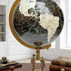 Könnte beinhalten: Ein dekorativer Globus mit einer schwarz-weißen Karte, montiert auf einem Messingständer mit Holzsockel. Der Globus wird von einem Messingmeridian getragen. Auf dem Tisch liegen Bücher und ein Tintenfass.