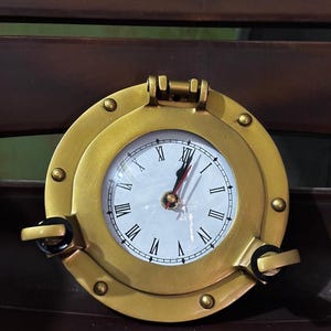 Könnte beinhalten: Eine runde Bullaugen-Uhr aus Messing mit weißem Zifferblatt, schwarzen römischen Ziffern sowie schwarzen Stunden- und Minutenzeigern und einem roten Sekundenzeiger. Der Rahmen hat ein oberes Scharnier und zwei seitliche Verschlussgriffe.