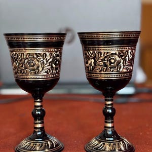 May include: Two black metal goblets featuring intricate gold-toned floral engravings around the upper bowl and base. Each chalice has a slender stem with horizontal gold bands and a flared rim, showcasing a contrasting dark and metallic finish.