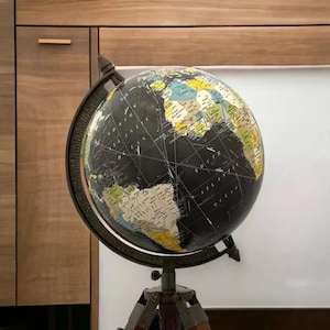 Peut inclure: Un globe terrestre noir et doré avec une carte détaillée, monté sur un support en métal foncé avec une base en trépied. Le globe présente une esthétique vintage, avec des continents en nuances de beige, vert et bleu.