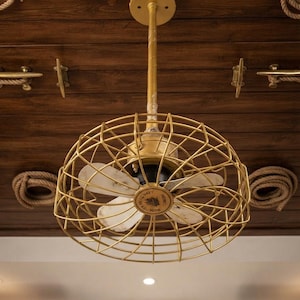 Puede incluir: Un ventilador de techo con una jaula de metal dorado y aspas blancas. El centro muestra el texto "ROYAL NAVY" y "MADE IN INDIA". El ventilador está suspendido por una varilla dorada sujeta a un soporte de techo circular.