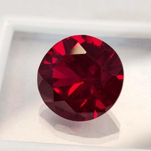 May include: A round, faceted gemstone with a deep, vibrant red color. The stone features a brilliant cut with numerous triangular and kite-shaped facets that reflect light, creating a complex pattern of dark and bright red tones across its surface.