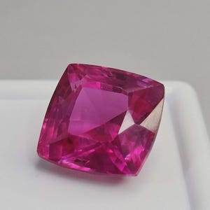 Puede incluir: Una gema vibrante, de talla cojín, con un tono magenta intenso. La superficie facetada refleja la luz, mostrando la claridad y el brillo de la gema. La forma cuadrada con esquinas redondeadas se presenta sobre un fondo blanco.