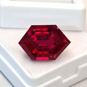 Op de afbeelding: Een gefacetteerde, zeshoekig geslepen, dieprode edelsteen. De edelsteen wordt gepresenteerd in een witte plastic behuizing, die zijn briljante facetten en rijke kleur laat zien. De vorm van de edelsteen is een gewijzigde zeshoek.