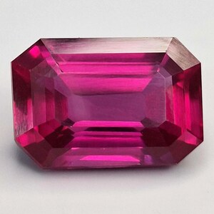 Peut inclure: Une pierre précieuse taillée en rectangle, d'un magenta vif. Les facettes de la gemme reflètent la lumière, créant un effet scintillant. La pierre est posée sur un fond blanc uni, mettant en valeur sa couleur et sa clarté. Idéale pour la joaillerie.
