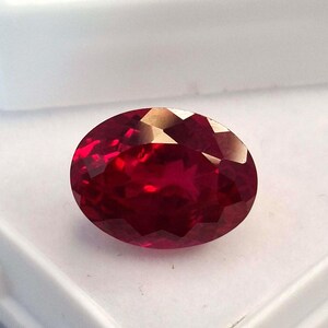 May include: A faceted, oval-shaped ruby gemstone. The gem is a deep red hue, with internal light reflections. The ruby is displayed against a white backdrop, accentuating its brilliance and clarity. The gemstone is the central focus, highlighting its natural beauty.