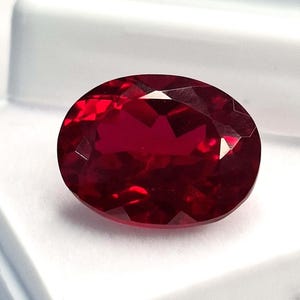 Peut inclure: Une pierre précieuse de type rubis, de forme ovale et facettée. La gemme est d'un rouge profond et saturé, avec de multiples facettes qui réfléchissent la lumière. Le rubis est posé sur un fond blanc.