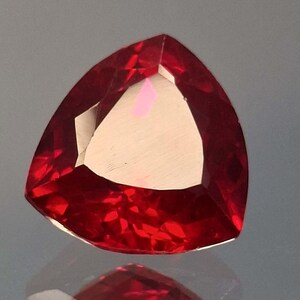 May include: A triangular-shaped, faceted gemstone in a deep red colour. The gem has a polished surface with multiple facets that reflect light, creating a brilliant sparkle. The gem's shape is a modified triangle, with a flat base and gently curved sides.