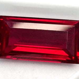 Peut inclure: Une pierre précieuse rectangulaire et facettée d'un rouge profond. La gemme a une apparence propre et polie, avec des bords nets et une forme symétrique. Le joyau est probablement un rubis, sur fond blanc.