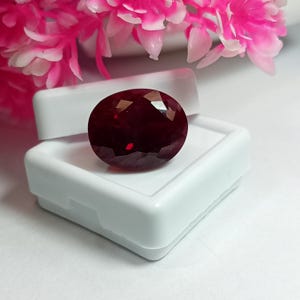 May include: A deep red, oval-cut gemstone with a faceted surface, displayed resting in a small, white, square plastic storage box. The gemstone exhibits a dark, saturated crimson hue with light reflecting off its polished, angular facets.