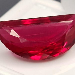 Op de afbeelding: Een gefacetteerde, halvemaanvormige edelsteen met een dieprode tint. De gepolijste oppervlakken van de edelsteen reflecteren licht, waardoor de interne structuur zichtbaar wordt. De steen staat tegen een witte achtergrond, wat de kleur en helderheid benadrukt. Geschikt voor sieraden.