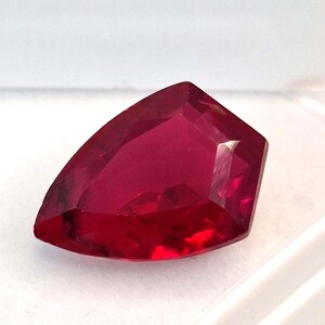 Könnte beinhalten: Ein facettierter, tiefroter Edelstein mit einer einzigartigen, eckigen Form. Der Edelstein hat einen hohen Reinheitsgrad und reflektiert das Licht, um brillante Facetten zu erzeugen. Der Stein ist vor einem weißen Hintergrund platziert, was seine satte Farbe und seinen Schliff hervorhebt.