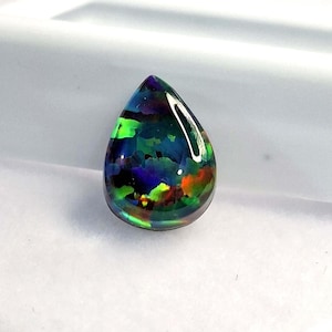 May include: A teardrop-shaped black opal gemstone with vibrant internal colors of green, blue, and red. The polished surface reflects light, showcasing the play-of-color effect. The gemstone is set against a white background.