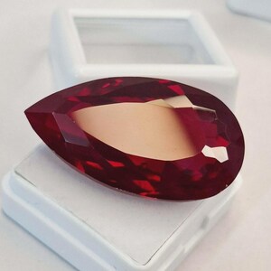 Op de afbeelding: Een gefacetteerde, druppelvormige edelsteen met een dieprode kleur en een lichtere, gouden binnenkant. De edelsteen wordt gepresenteerd op een witte, vierkante basis, wat duidt op een presentatie voor sieraden of verzamelobjecten. De slijping creëert meerdere reflecterende oppervlakken.