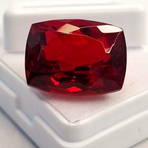 Puede incluir: Una gema facetada, tallada en cojín, de color rojo intenso. La gema se presenta sobre un fondo blanco, resaltando su claridad y brillo. El corte de la piedra crea múltiples superficies reflectantes.