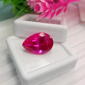 May include: A pear-shaped, faceted gemstone in a vibrant, deep pink color. The stone is displayed resting on the white plastic base of an open jewelry box, with its polished surfaces reflecting light to highlight the intricate cut and clarity.