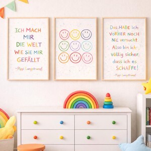 May include: Three framed art prints with colourful text and smiley faces hang on a white wall. The prints feature inspirational quotes in German. A white dresser with colourful knobs and a rainbow decor are in front of the wall.