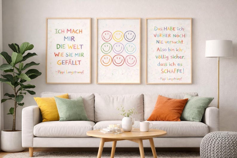 May include: Three framed art prints with German text and smiley faces in rainbow colours. The prints are displayed above a white sofa with colourful cushions. A round wooden coffee table and a potted plant complete the room's decor.