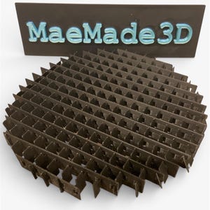May include: A brown, circular 3D-printed object with a honeycomb-like structure. The object is in front of a dark brown sign with the text "MaeMade3D" in light blue, 3D-printed letters. The background is white.