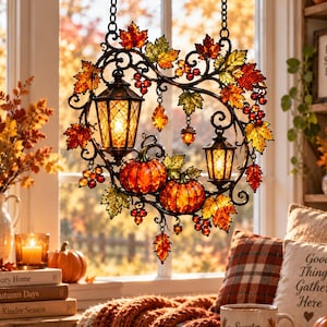 Op de afbeelding: Een decoratief hangend ornament met twee verlichte lantaarns, twee oranje pompoenen en een krans van herfstbladeren in rood-, geel- en groentinten. Het metalen frame is versierd met kleine bessen, eikels en bungelende kristallen accenten.