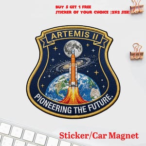 May include: A navy blue Artemis II patch with gold trim, featuring a rocket launching from Earth towards the moon. The patch includes the text "ARTEMIS II" and "PIONEERING THE FUTURE." The image also includes the text "BUY 5 GET 1 FREE STICKER OF YOUR CHOICE [2X2 SIZE]" and "Sticker/Car Magnet."