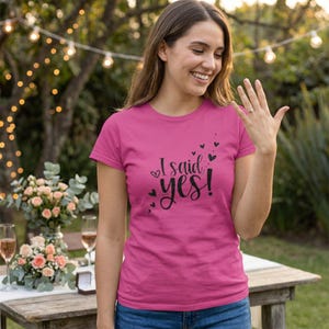 Può includere: Una maglietta magenta con la scritta "I said yes!" in nero e cuori decorativi. La donna sorride e mostra un anello sulla mano sinistra. Lo sfondo include un tavolo con fiori e bicchieri.