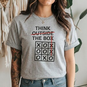 May include: A gray t-shirt with the text "THINK OUTSIDE THE BOX" in black, with the word "OUTSIDE" crossed out in red. Below the text is a tic-tac-toe board with X's and O's. The shirt is a casual style.