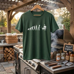 May include: A dark green t-shirt with a white graphic depicting the evolution of grilling. The graphic shows a progression from ape to chef at a grill. The shirt is hanging on a wooden hanger in an outdoor setting with a grill and accessories.