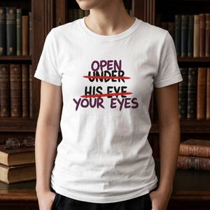 Può includere: T-shirt bianca con la scritta "OPEN UNDER HIS EYE YOUR EYES" in viola, con le parole "UNDER" e "HIS EYE" sbarrate in rosso. La maglietta è indossata davanti a una libreria.