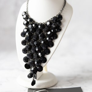 Könnte beinhalten: Eine schwarze Perlenkette mit einem kaskadierenden Design. Die Kette wird auf einem weißen Ständer präsentiert. Der Text "CagnetteGirls Creative Jewellery" ist auf einer Karte unter dem Ständer sichtbar.