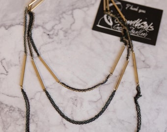 Puede incluir: Un collar de capas con tres cadenas de barra dorada y dos cadenas negras. El collar está sobre una superficie de mármol blanco. El texto "Sweetgirls Creative Jewellery" está en una etiqueta negra.
