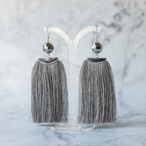 May include: A pair of silver earrings with gray tassel fringe. The earrings feature a round, faceted silver bead at the top of each tassel.