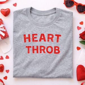 May include: A heather gray t-shirt with the words "HEART THROB" printed in distressed red lettering. The shirt is surrounded by Valentine's Day themed decorations, including red heart-shaped sunglasses, hearts, and a red rose.