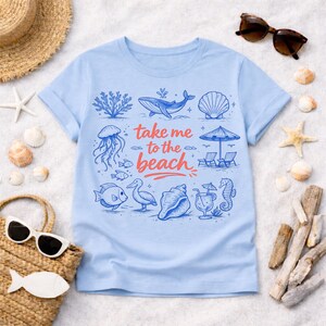 K&ouml;nnte beinhalten: Hellblaues T-Shirt mit einem Strandmotiv. Das Design zeigt Illustrationen eines Wals, einer Qualle, Fischen und Muscheln sowie den Text "take me to the beach" in Rot und Blau. Das T-Shirt ist von Strandaccessoires umgeben.