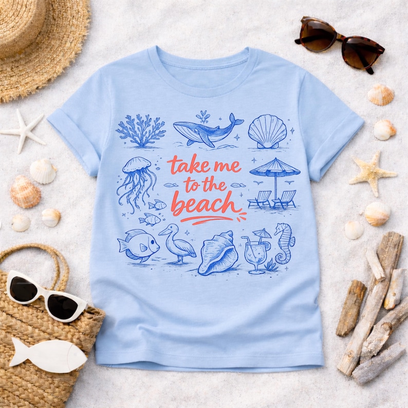 K&ouml;nnte beinhalten: Hellblaues T-Shirt mit einem Strandmotiv. Das Design zeigt Illustrationen eines Wals, einer Qualle, Fischen und Muscheln sowie den Text "take me to the beach" in Rot und Blau. Das T-Shirt ist von Strandaccessoires umgeben.