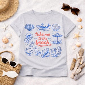 K&ouml;nnte beinhalten: Hellgraues T-Shirt mit Strandmotiv. Das Design umfasst blaue Illustrationen von Meereslebewesen und Strandelementen, wie einem Wal, einer Qualle und Muscheln. Der Text "take me to the beach" ist in Rot und Blau gehalten. Sonnenbrille und Strohhut sind sichtbar.