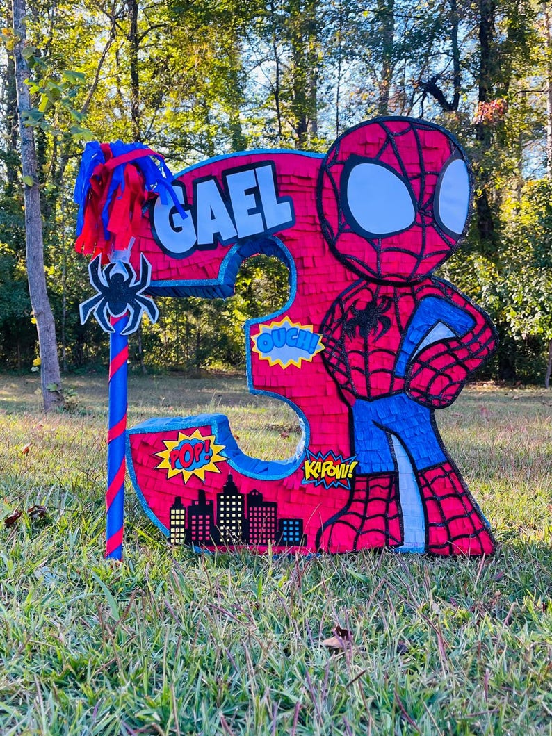 May include: A large, handmade pi&ntilde;ata in the shape of the number 3, featuring a Spider-Man theme. The pi&ntilde;ata is primarily red and blue, with the name "GAEL" displayed. Comic book sound effects and a cityscape are also included.