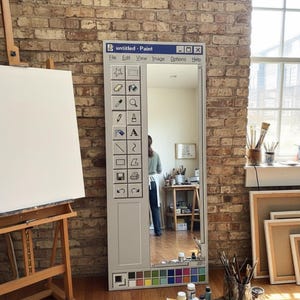 Può includere: Uno specchio a figura intera progettato per assomigliare a un'interfaccia di programma di pittura per computer, con una tavolozza di colori in basso. Un cavalletto da artista con una tela bianca è a sinistra. Materiali per la pittura e tele incorniciate sul pavimento.