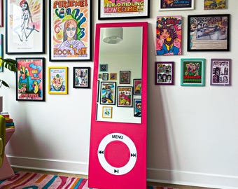 May include: A vibrant pink full-length mirror designed to resemble a classic digital music player. The mirror features a white circular control panel with directional arrows and a "MENU" button. The wall behind the mirror is adorned with framed artwork, and a colorful rug is visible.