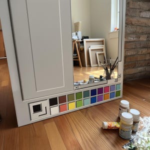 May include: A gray cabinet with a mirror and a built-in color palette. The palette features a variety of colors, including black, white, red, yellow, green, blue, and purple. Paint tubes and jars are on the floor.
